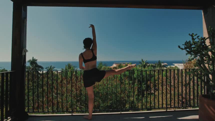 A woman practices a graceful yoga pose on a sundrenched balcony resting her leg on the railing while overlooking a lush tropical landscape and the sparkling blue ocean