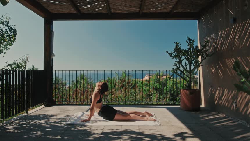 Young adult female doing yoga exercises on top of a mat with a balcony with a natural landscape on the background during day time