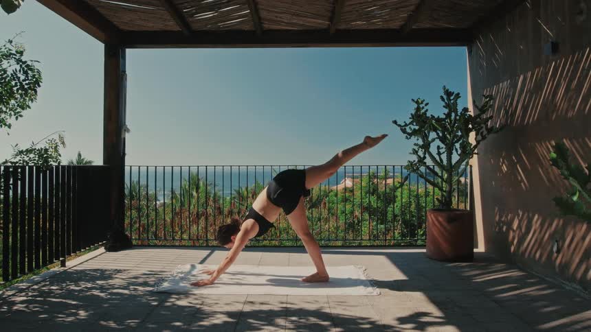A woman in a black sports top and shorts performs a onelegged yoga pose on a white mat under a wooden pergola looking forward with a focused and serene mood