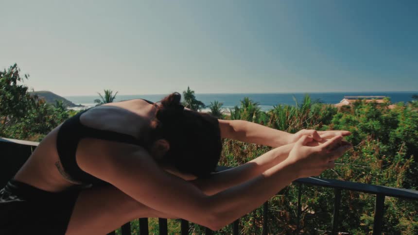 A young woman in athletic wear stretches her leg on a balcony railing while overlooking a lush tropical landscape and the ocean under a clear blue sky