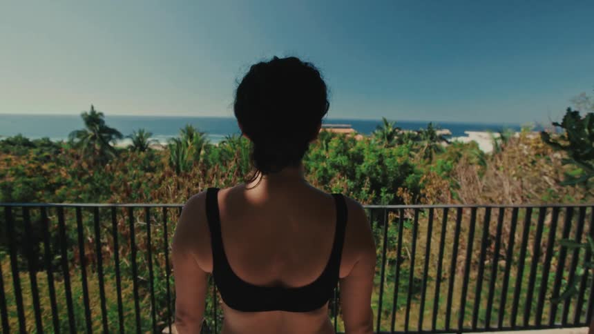 Woman standing with her back on a balcony overlooking vegetation and the ocean