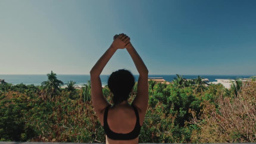 A woman stands with arms lifted on a balcony overlooking a tropical coastline in daylight