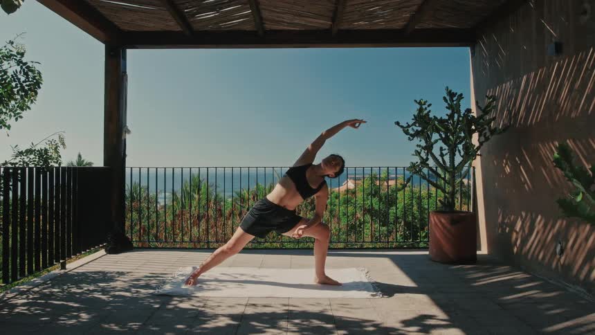 A woman practices a yoga warrior pose on a covered terrace overlooking the ocean in daylight