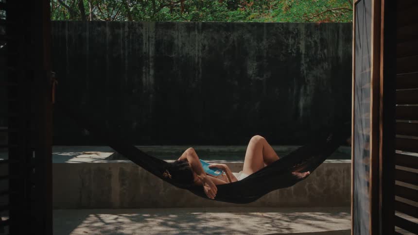 Woman lying in a hammock outdoors holding a book in a shaded area