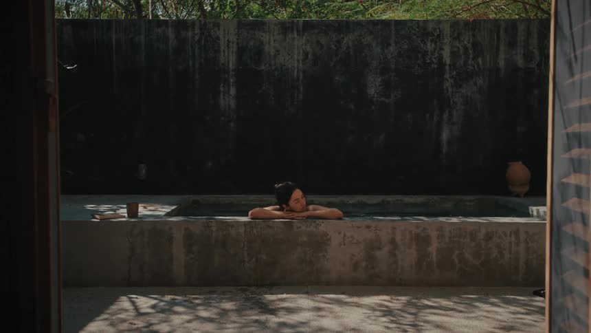A young woman relaxes in a small rectangular concrete pool set against a high dark wall in a sunlit courtyard