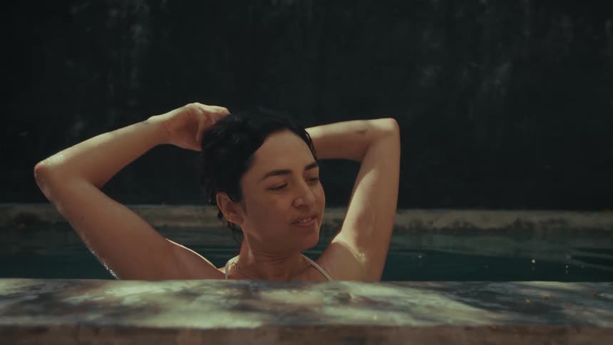 A woman with dark hair stands in a stone pool raising her arms to gather and adjust her hair while looking off to the side with a calm expression against a dark textured wall