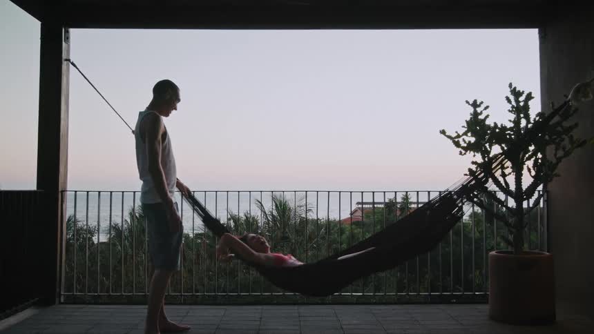 Man swings a woman lying in a hammock on a balcony overlooking the sea at sunset