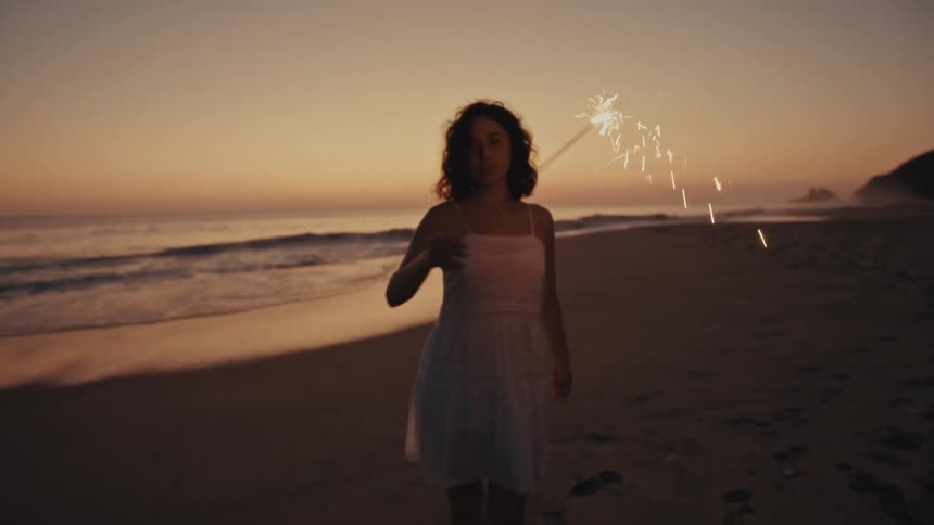 A woman in a white dress dances on a beach at sunset while holding a lit sparkler that trails sparks through the air