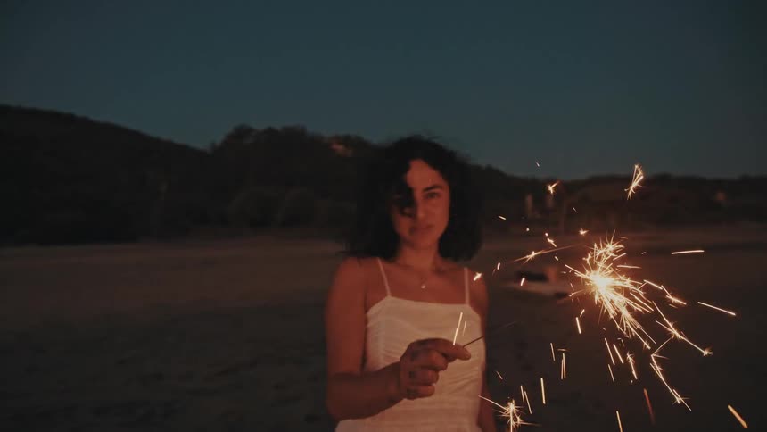 A woman joyfully plays with a sparkler on the sandy beach at dusk near the ocean