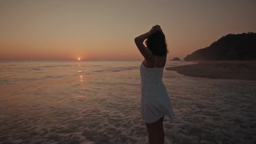 A woman in a white dress stands in the shallow surf on a beach while looking out at the sunset over the ocean