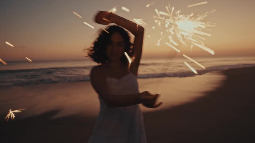 A woman joyfully dances along the shoreline at sunset waving a brilliant sparkler that casts glowing sparks against the warm twilight sky