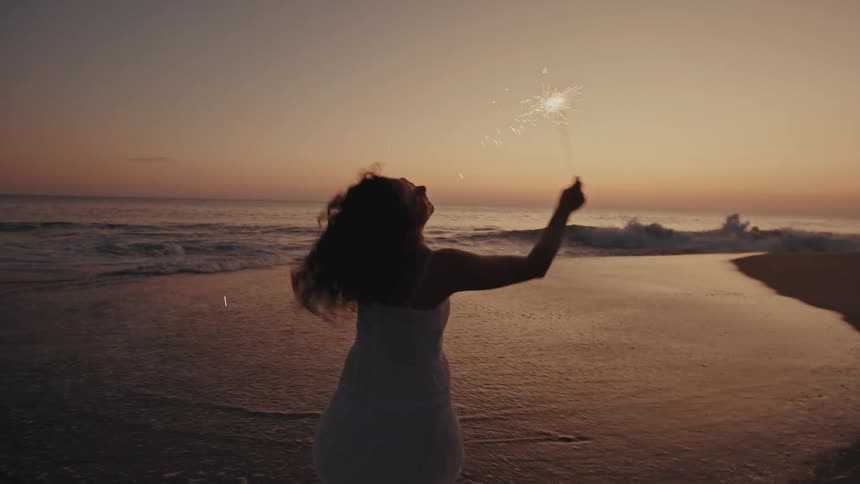 A woman with dark curly hair wearing a white dress dances and twirls on a sandy beach while holding a sparkling firework at sunset