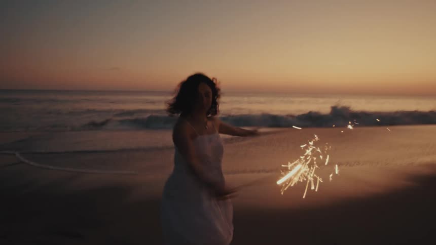 Woman twirls on the beach in the early evening while holding a sparkler