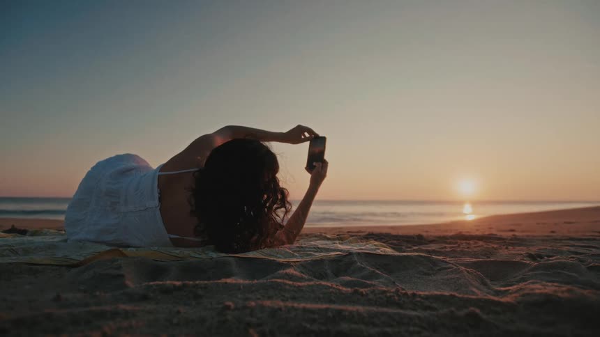 The woman takes a photo of the rising sun with her cell phone on the beach