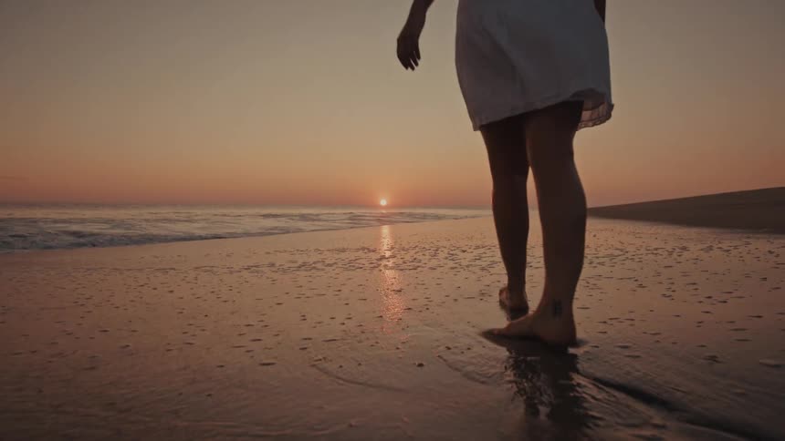 From a low angle the camera follows a womans bare feet as she strolls along the wet shoreline the golden sunset reflecting on the receding waves