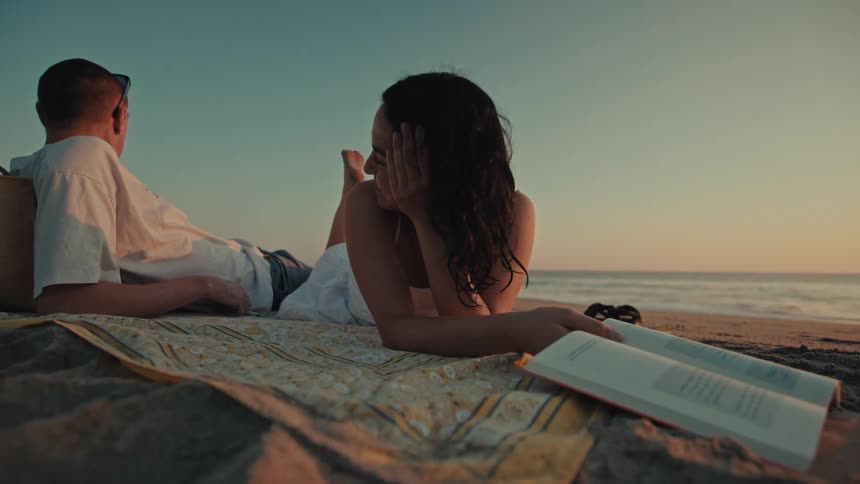 A couple lies on a beach blanket having a relaxed conversation by the ocean at sunset