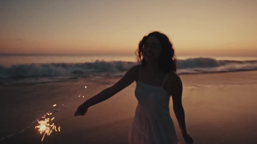 A young woman dances on the beach at sunset holding a lit firework that sparks and glows as the sky turns warm and colorful