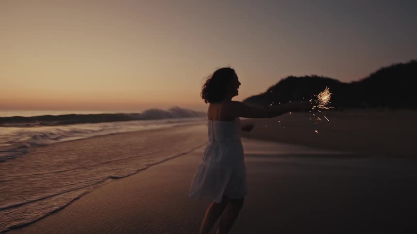 Teenage girl in a white dress running on the beach with a sparkler at dusk