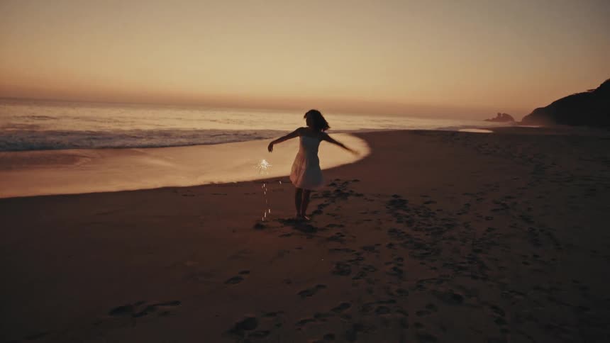 Woman walking along the beach at sunset with arm raised near the sea