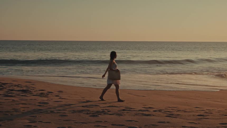 Young woman with a large handbag walks across the beach as waves roll in to shore