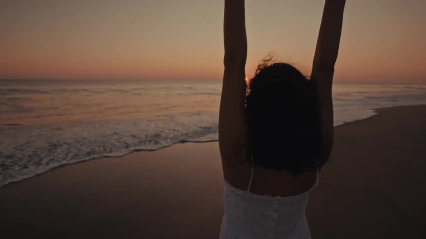 Silhouetted against a glowing sunset a woman stands on the wet sand of a beach and slowly raises her arms to the sky embracing the tranquil moment as waves gently wash ashore