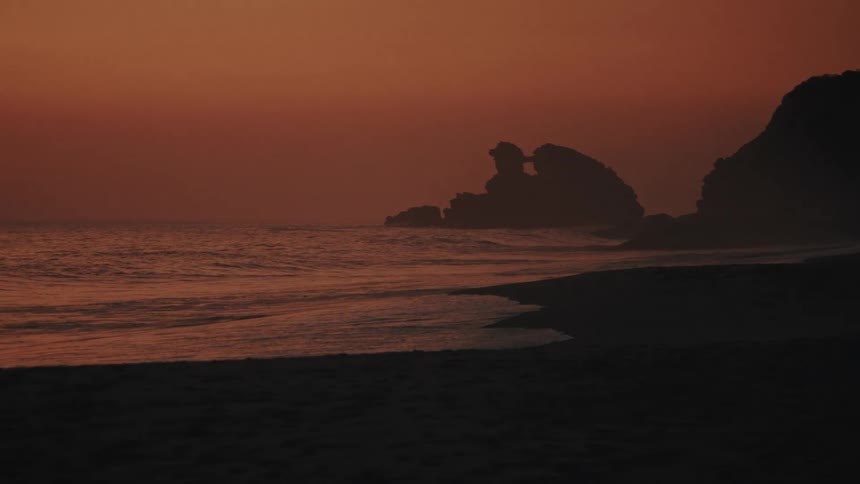 A deep orange sky glows after sunset as waves wash over a darkened beach outlining the dramatic silhouettes of a rocky coastline in the distance