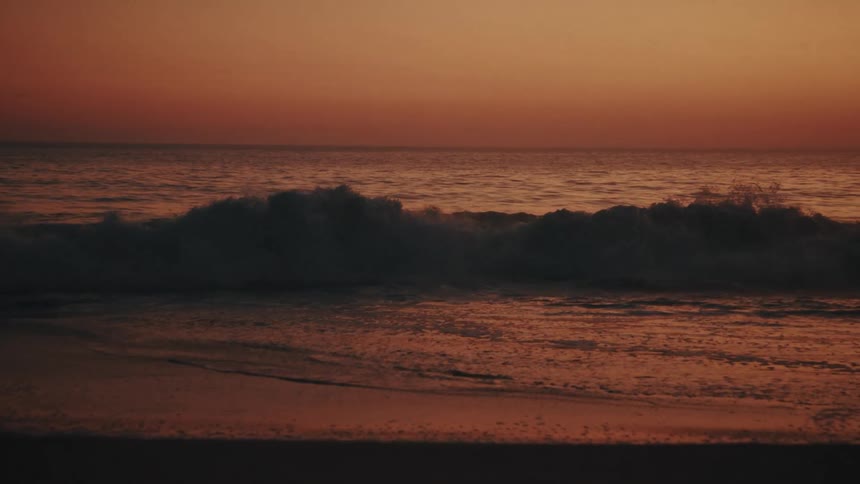 Small waves crest and crash against the shore of a dark beach at sunset with the deep orange glow of the sky reflecting off the moving water and damp sand