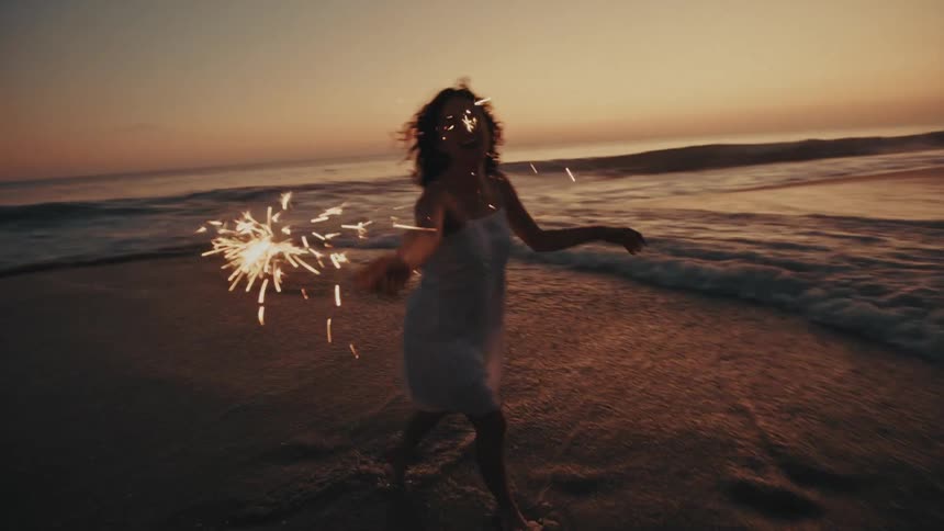 Woman laughs and wades through the surf while holding a lit sparkler in the evening