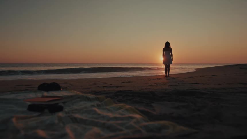 Silhouette of a young adult woman from the bacl wearing a white dress standing on a beach near the ocean outdoors during dusk or dawn