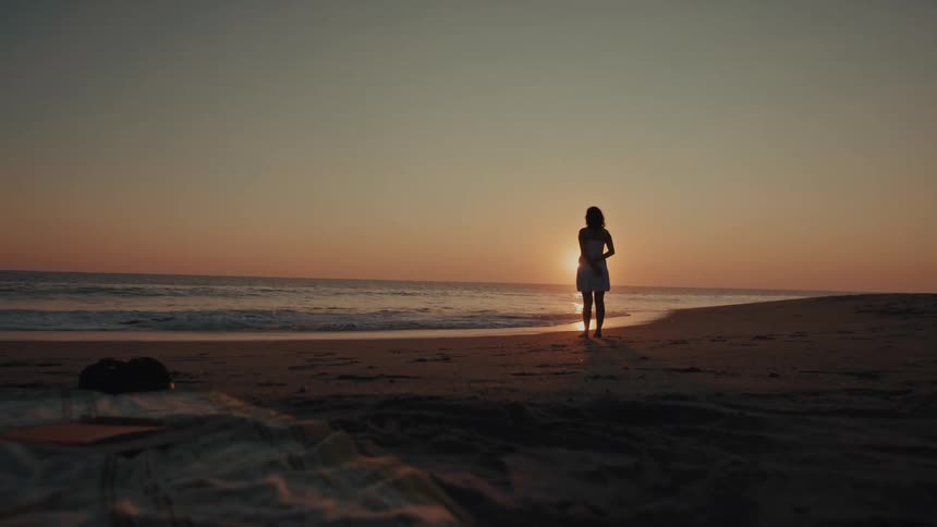 Teenage girl in a white dress stands on the beach as waves roll in to shore and the sun glows on the horizon