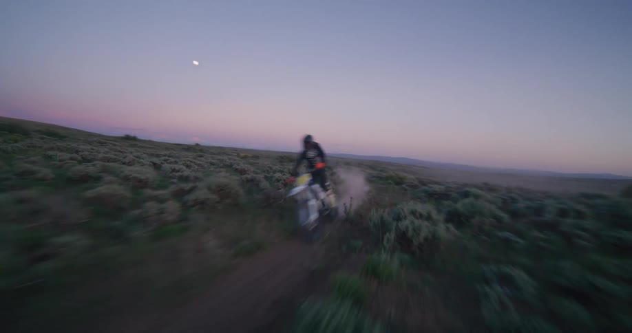 Biker riding a motorcycle on a dust road surrounded by vegetation with hills on the background during dusk or dawn