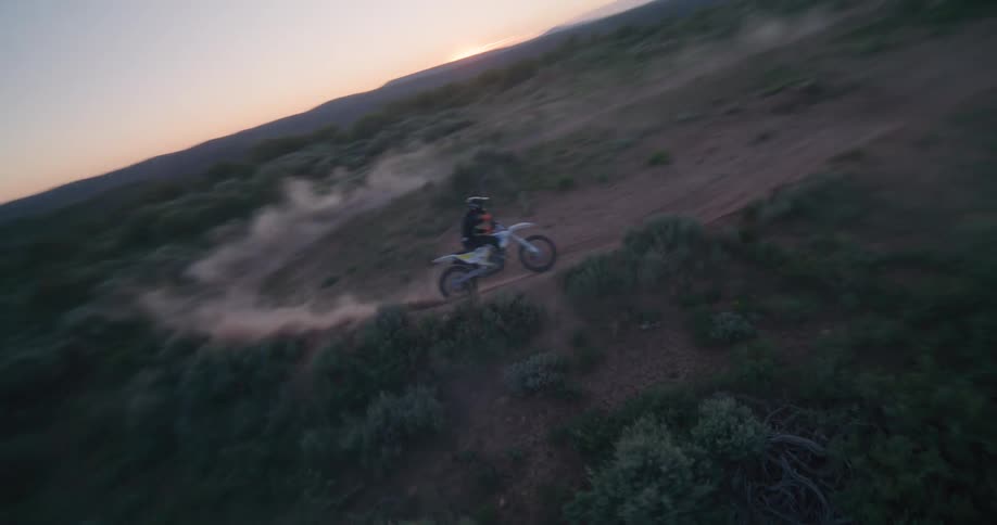 A person rides a motocross bike at high speed along a winding dirt track through a desert landscape at dusk kicking up a trail of dust as he navigate the terrain under a fading sunset sky