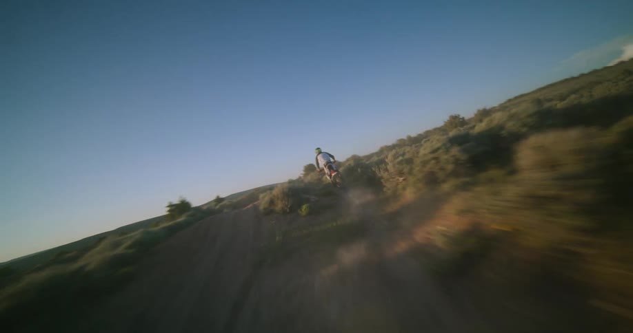 A dirt biker speeds down a dusty mountain trail during a bright sunset kicking up dirt as the camera follows closely from behind to capture the fastpaced offroad action through the desert shrubland