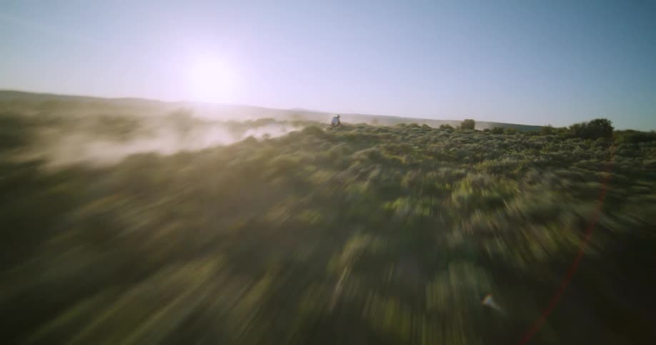 A person rides a dirt bike down a dusty trail on a steep brushcovered hillside during a sunny day with the camera following closely behind to capture the speed and the cloud of dust kicked up by the tires