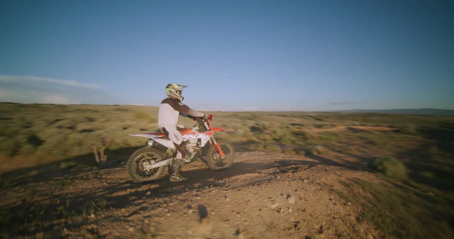 A mountain biker stands on top of a hill fully kitted in a jacket and helmet then launches forward and speeds down the slope with the setting sun glowing behind him