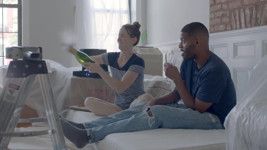 A man and woman drink champagne while sitting on a mattress on the floor