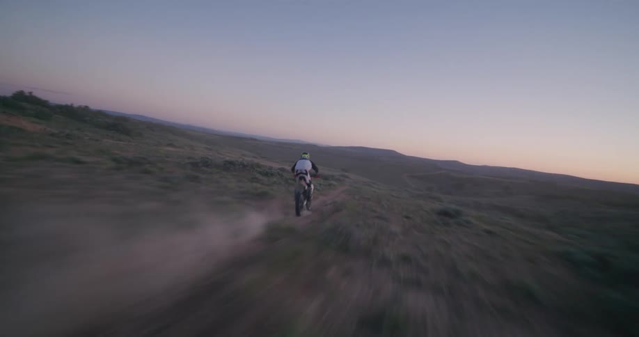 A motorcyclist speeds across a dusty desert trail at sunset in a vast landscape