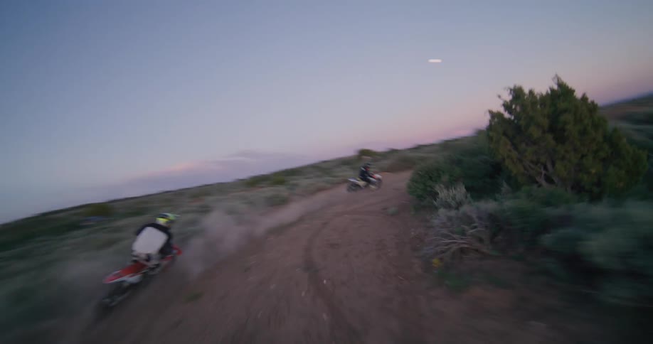 An fpv drone follows two motocross riders as they speed along a winding dirt track performing jumps and kicking up dust in a vast open landscape at dusk