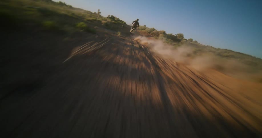 A person is riding a motorcycle down a dirt path through a grassy field under a clear blue sky looking forward with a sense of adventure