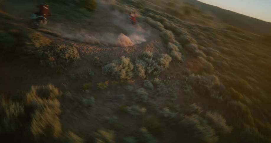 Two dirt bikers race across a dusty desert trail at sunset kicking up clouds of sand as they navigate turns and small jumps through the scrubfilled landscape under a bright sun
