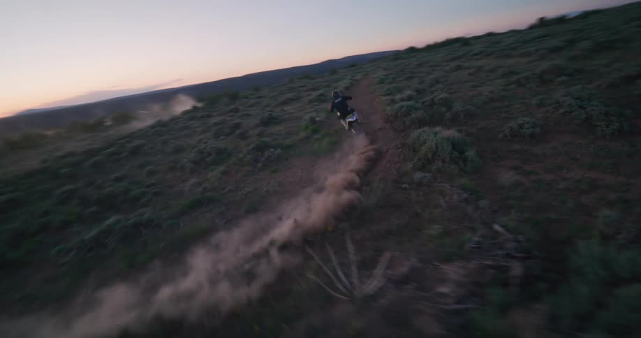 A motorcyclist rides across open scrubland at dusk kicking up dust under a fading sky