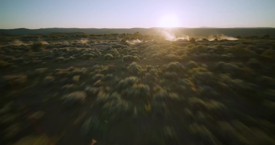 Aerial drone view flying low over two motorcyclists jumping on a dusty ramp on a vast green plain toward a bright sun on the horizon during a golden sunset