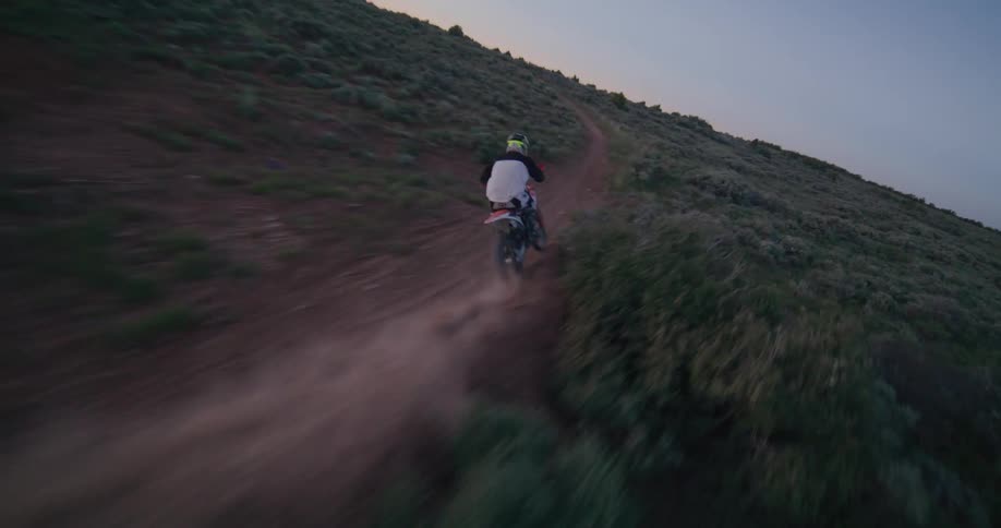 An fpv drone follows a motocross rider as they navigate a winding dirt track through a brush covered landscape at sunset