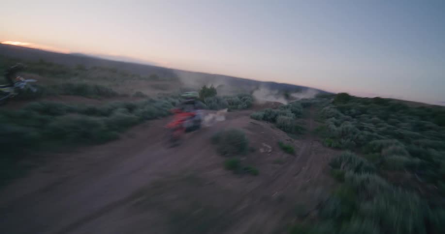 An fpv drone follows a motocross rider as they navigate a winding dirt track through a brush covered landscape at sunset