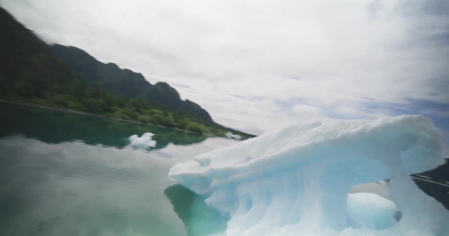 An fpv drone captures a dynamic view flying over and around several small icebergs floating in a calm bay surrounded by lush green mountains and snowy peaks