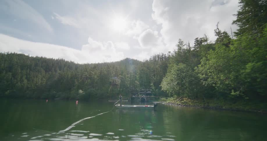 A distant view shows a person fly fishing from a small boat on a calm lake surrounded by a dense evergreen forest under a bright cloudy sky