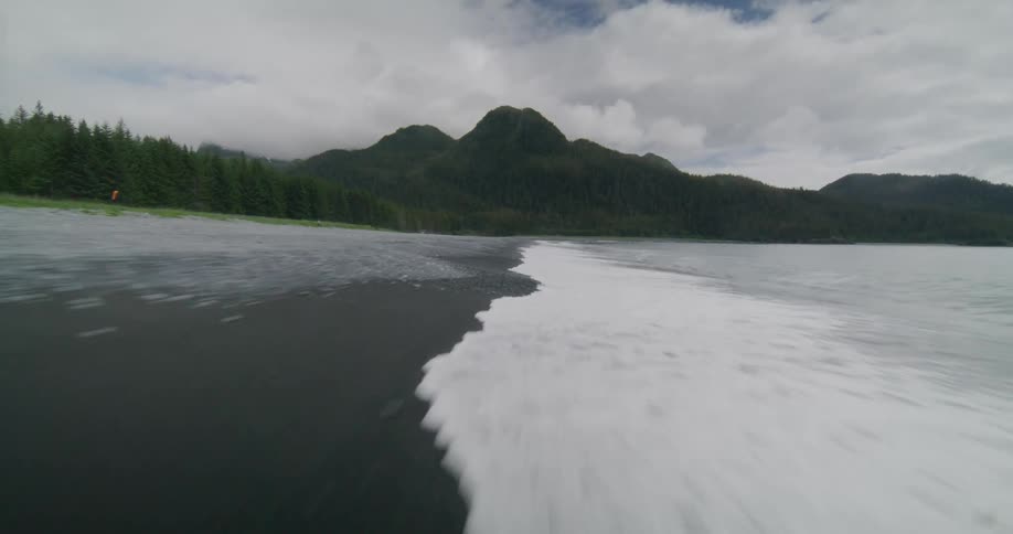 An aerial view speeds low along a black sand beach towards a forested mountain