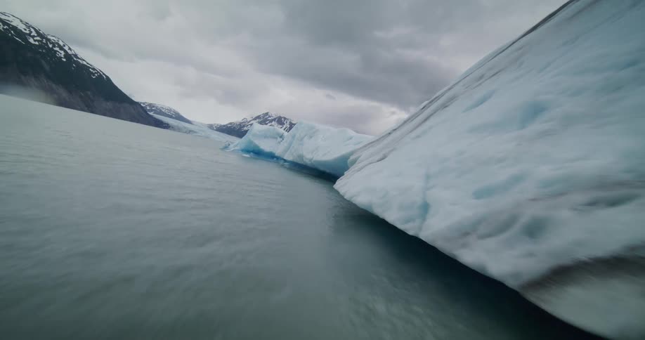 An iceberg floats on a lake bordered by a mountain range their peaks covered in snow beneath a cloudy sky