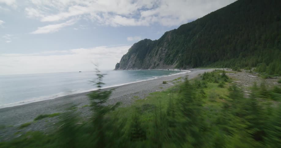 An island with a tall mountainous hill covered in trees as waves crash against the shoreline at midday