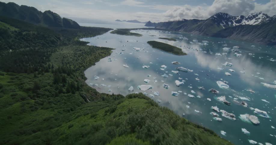 An iceberg floats on a lake at midday surrounded by a mountain range the distant peaks covered in snow while the mountains in the foreground are lined with green vegetation
